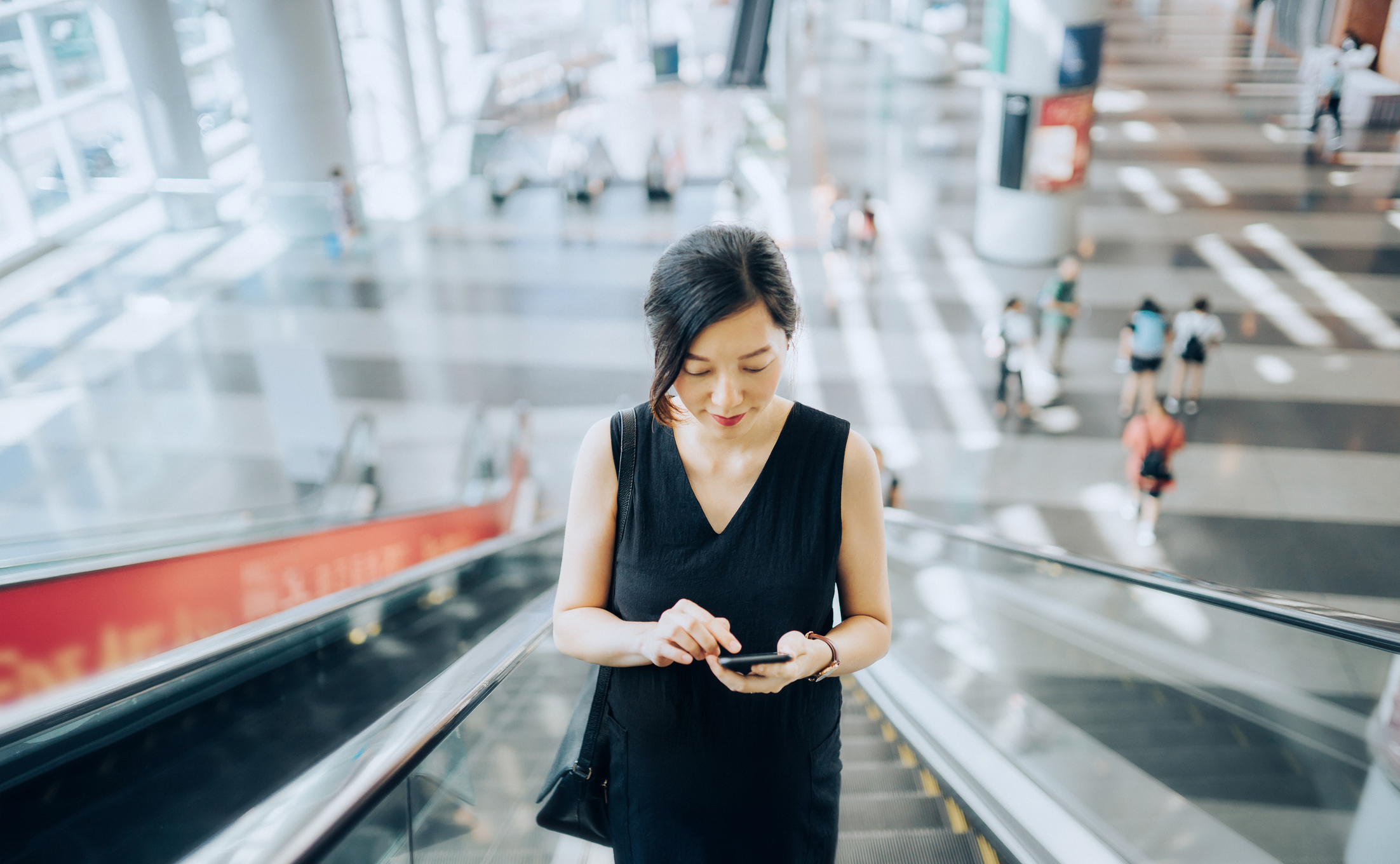 Young businesswoman reading emails on smartphone while riding on escalator.